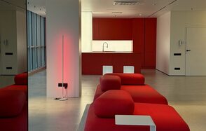 Арт-Плейс Фактура. Современный лофт с видом на весь город. Современное пространство с красной мебелью, кухонной зоной с красными шкафами и белой раковиной. Большие окна по одной стене, пол из светлого дерева или плитки. Вертикальный светильник у стены. Белые столики. Стены светлые, одна дверь.