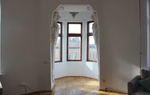 Cassiopea(Кассиопея). Эркер с большими окнами, деревянный паркет, светло-белые стены, барельефы с фигурным декором, потолочный светильник, частично виден диван с подушками.