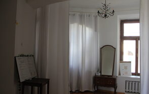 Cassiopea(Кассиопея). Комната с белыми стенами и шторами. В центре зеркало на деревянном комоде. Правее окно с белой рамой и радиатором. На комоде картины в рамках. В левом углу стоит стол с черной доской. Потолок оформлен черной люстрой.