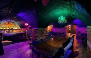 GB Rooms(ГБ Румс). Лофт №1. Кирпичное подвальное помещение с арочным потолком. Длинный деревянный стол, окружённый стульями. Барная стойка на заднем плане. Зелёная и фиолетовая подсветка. Классическая люстра. Пол деревянный. Мягкая мебель вдоль стен.