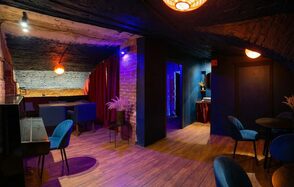 GB Rooms(ГБ Румс). Лофт №1. Подвальное помещение с кирпичными стенами и деревянным полом. Зоны с мягкими синими креслами и диванами, столики. Яркое цветное освещение, красные шторы. Потолок темного цвета. Круглые лампы с теплым светом.