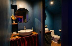 GB Rooms(ГБ Румс). Лофт №1. Ванная комната с синими стенами, круглым зеркалом над раковиной, круглой раковиной на деревянной столешнице, унитазом. Металлический дозатор мыла, красная занавеска под столешницей. Полки с декором, верхнее точечное освещение.