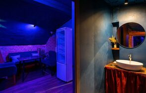 GB Rooms(ГБ Румс). Лофт №1. Комната с синим и красным освещением, кирпичные стены, черная мебель, обеденный стол, диваны, холодильник, круглая раковина с зеркалом, деревянный и керамический декор, занавеска красного цвета, темный деревянный пол.