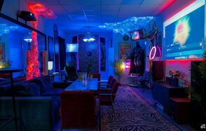 GB Rooms(ГБ Румс). Лофт №2. Квартира с затемнённым освещением, столом и креслами, диванами, неоновыми вывесками и декоративными элементами. На стенах постеры, есть крупный экран для проекций. Используются яркие синие и красные цветовые акценты. На полу ковер с узором.