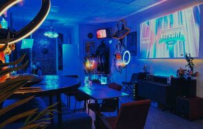 GB Rooms(ГБ Румс). Лофт №2. Комната с неоновым освещением, большой стол и стулья, диваны, проектор с изображением на стене, тематические постеры и декоративные элементы. Стены светлые, пол паркетный, на некоторых поверхностях светящиеся неоновые элементы.