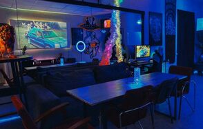 GB Rooms(ГБ Румс). Лофт №2. Помещение для отдыха с неоновым освещением в синих тонах. Длинный стол с несколькими стульями, мягкое сиденье, телевизор, настенные картины. Пламя в центре стола. Просторная обстановка с тёмной мебелью.