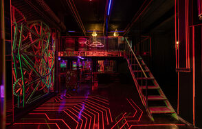 Лофт Космос. Зал Ночной Клуб. Темное помещение с черным потолком и стенами, разноуровневая планировка с лестницей. Неоновая подсветка красного и синего цветов на полу и стенах создает геометрические узоры. Металлические конструкции, подвесные лампы.