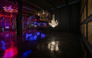 Лофт Космос. Зал Ночной Клуб. Просторное помещение с бетонным полом и черными стенами. Лестница с прозрачными перилами ведет на второй уровень. Неоновая подсветка, люстра и несколько висящих ламп освещают пространство. Часть стены закрыта плотной черной занавеской.