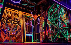 Лофт Космос. Зал Ночной Клуб. Праздничное пространство с ярким неоновым декором, включающим геометрические узоры на стенах и конструкциях. Преобладающие цвета: оранжевый, зеленый и фиолетовый. Подвесное освещение и элементы напоминающие механизмы, надпись "ONSPACE".