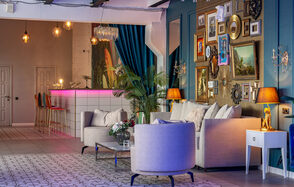 Лофт Май. Стильное пространство с верандой. Гостиная с баром в современном стиле. Белые диваны и кресла, стол с цветами. На стене множество картин и зеркал в рамах. Бар с высокими стульями и розовой подсветкой. Потолочные лампы, декоративные растения, синие шторы. Стены бирюзовые, полы плиточные.