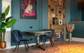 Лофт Май. Стильное пространство с верандой. Угловое пространство с деревянным круглым столом и двумя синими стульями. На стене синие панели, висят картины и фотографии. Полка с декором и несколькими книжными полками. На полу плитка с узором, рядом растение в горшке.