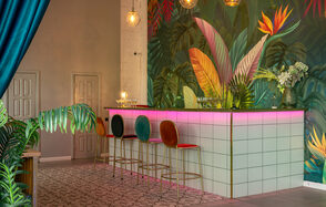 Лофт Май. Стильное пространство с верандой. Барная стойка с белыми квадратными плитками, подсветка розового цвета. Три барных стула с разноцветными спинками. Стена с ярким тропическим рисунком. Зона декорирована зелеными растениями, висят круглые подвесные светильники.