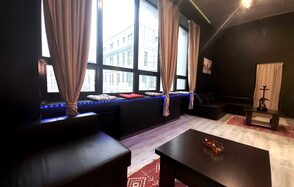 Элегантный лофт с хорошим звуком и балконом. Лофт с чёрными стенами и большими окнами, с бежевыми шторами. Низкие чёрные кожаные диваны и деревянные столы. Красные ковры на светлом полу. На одном из столов расположен кальян. Искусственное освещение, вид на город.