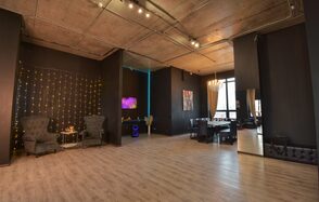 Элегантный лофт с хорошим звуком и балконом. Просторное помещение с высокими потолками и деревянным полом. Черные стены, на одной из которых гирлянда. Зона с креслами и столиком, рядом телевизор. Обеденная зона с большим столом и стульями. Потолочные светильники и люстра. Большие окна с занавесками.