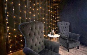 Элегантный лофт с хорошим звуком и балконом. Тёмно-серая комната с двумя мягкими креслами, круглым белым столиком, на котором стоит декоративный светильник. На стене гирлянда из маленьких жёлтых огней. Пол из светлого дерева.