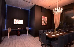 Элегантный лофт с хорошим звуком и балконом. Пространство для мероприятий с деревянным столом и синими стульями, сервированное столовыми приборами и бокалами. Черные стены, подсветка по периметру одной стены, телевизор, картина. Люстра с лампами, светлая штора, декоративные элементы.