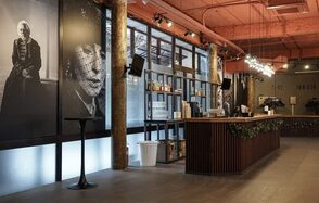 Театр в Хамовниках. Барная зона в просторном помещении с высокими потолками. Стойка из темного дерева с растительной декорацией. Стены украшены крупными черно-белыми портретами. Освещение подвесное, имеется множество бутылок. Потолок красный.