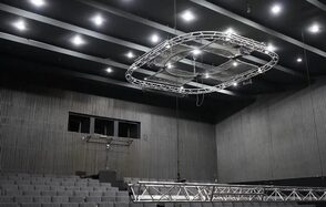 Театр в Хамовниках. Театральное или конференц-пространство с амфитеатром, оборудовано рядами серых кресел. Потолок черный с вмонтированными светильниками и подвесной конструкцией. Стены облицованы темным древесным материалом.