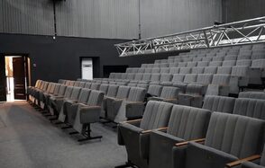 Театр в Хамовниках. Аудитория с рядовой расстановкой серых кресел. Настенное покрытие и пол темные. Освещение отсутствует, видны металлические конструкции для светового оборудования. Открытая дверь на заднем плане.