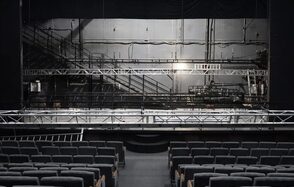 Театр в Хамовниках. Театральный зал с рядами серых кресел и сценой. На сцене установлены металлические конструкции и оборудование. Задняя стена сцены с проводами и аппаратурой. Освещение верхнее, стены серые.