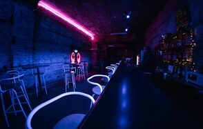 PVZ CLUB(ПВЗ Клуб). Барное пространство с высоким потолком и кирпичными стенами, обставлено высокими столами и стульями. Неоновая подсветка фиолетового цвета, барная стойка справа. На стенах полки с напитками, справа заметны гирлянды.