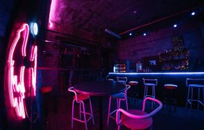 PVZ CLUB(ПВЗ Клуб). Тёмное помещение с барной стойкой, окруженной высокими стульями с красными сиденьями. Справа неоновые огни и вывеска на стене. Кирпичные стены, деревянный пол. Освещение синих и фиолетовых оттенков. На барной стойке видны напитки и декорации.