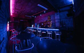 PVZ CLUB(ПВЗ Клуб). Темное индустриальное пространство с кирпичными стенами и бетонным потолком. Барная стойка с высоким освещением и стульями. Несколько круглых столов и стульев с белыми рамами и красными сиденьями. Неоновая подсветка в фиолетовых и синих тонах.