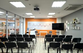 Конференц-зал. Конференц-зал с креслами на несколько рядов, бело-оранжевая стена с надписями, большая плоская панель на подставке, стеклянная перегородка слева, светлые стены и пол, потолочные светильники, кондиционер, полки с наградами и сертификатами.