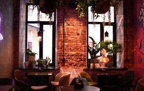 Винный бар “Дело не в вине”. Лофт-пространство с кирпичными стенами и высокими окнами. Разнообразные кресла и столы, декоративные растения и бюст. Подвесные светильники обеспечивают мягкое освещение. Яркий, уютный интерьер с граффити на стенах и неоновыми акцентами.