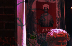 Винный бар “Дело не в вине”. Темное арт-пространство с кирпичной стеной, картина в рамке с изображением человека в шляпе на красном фоне, гипсовая скульптура и растение на переднем плане, цветное освещение.