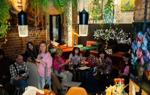 Винный бар “Дело не в вине”. Кафе с кирпичными стенами, мягкими скамейками, деревянными столами и стульями. Потолочные светильники, зелёные растения, яркие картины и граффити. Посетители за столами с напитками.