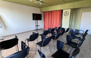 Первый План. Аудиторное пространство с рядами черных стульев, телевизором на стойке, красной занавеской, потолочными светильниками и настенными часами. Пол деревянный, стены светлые, частично индустриального стиля.