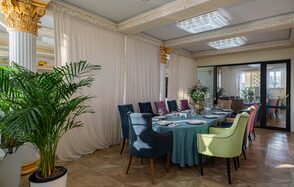 Вип-зал. Элегантный банкетный зал с круглым столом, окружённым разноцветными стульями. На столе сервировка из тарелок и бокалов. Помещение украшено колоннами с золотыми деталями, зеркалами, растениями в горшках, подвесным потолком с квадратными светильниками.