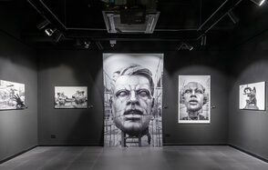 FOTOLAB(ФОТОЛАБ). Выставочное пространство с черными стенами и потолком. Черно-белые фотографии на стенах с изображениями лиц и сцен. Освещение точечное, подвесное. Пол из темного плиточного материала. Центральная картина с крупным изображением лица.