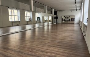 The Flow Space(Пространство Поток). Просторное помещение с деревянным полом, зеркальными стенами и большими окнами. Профессиональное освещение на потолке, колонки на стойках в углу. Белые стены и потолок, несколько батарей под окнами.