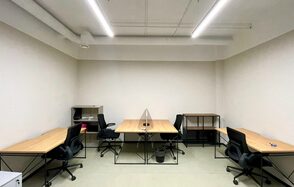 Smart Coworking(Умный Коворкинг). Офисное пространство с пятью рабочими местами, каждое с деревянным столом и черным офисным креслом. Белые стены, белый потолок с трубами и подвесным длинным освещением. Пол светлый, на столах и полу несколько предметов.