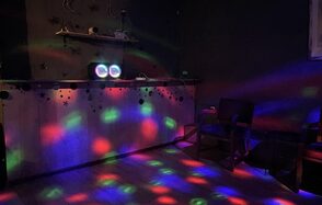 Лофт ВЕЧЕ’Р. Тёмное помещение с деревянным полом, барная стойка, украшенная светящимися гирляндами. Яркие разноцветные световые болты на стенах и полу. Стена украшена полкой с декоративными элементами. Две деревянные кресла сбоку.