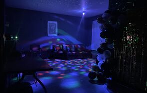 Лофт ВЕЧЕ’Р. Тёмная комната с диванами и креслами, подсвеченная разноцветными световыми эффектами. Стены тёмные, на одной из них картина. В правом углу арка из воздушных шаров. Поликающее освещение создаёт атмосферу вечеринки.