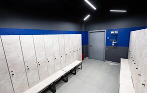 Футбольный манеж “РФЛ-Арена” имени летчика-космонавта А. А. Леонова. Гардеробная комната с серыми шкафчиками и скамейками. Светлые стены и плиточный пол. Синий цвет в верхней части стен. Светодиодное освещение на потолке. Закрытая дверь и настенное оборудование.