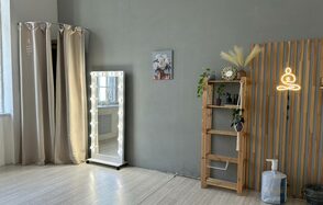 Лофт СВЕТОСФЕРА. Просторная комната с серыми стенами и деревянным полом, зеркало с подсветкой, светлая шторка в углу, деревянная полка с растениями и декором, настенный постер, неоновая лампа в виде сидящего человека, плотные шторы, серое кресло.