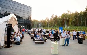 Технопарк «Университетский». Открытая площадка.. Открытое пространство с деревянными диванами и столами, белая тентовая конструкция, металлические бочки используются как высокие столы, панорамное стеклянное здание на заднем плане, вечернее освещение, зелёные деревья вокруг.