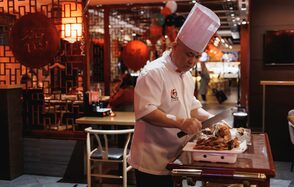 Ресторан J.Z.Peking Duck (J.Z. Утка по-пекински) . Ресторанное пространство, повар в форме режет мясо на передвижном столике. Слева решетчатая перегородка, декорированная красным кругом. Интерьер в тёплых тонах, видны красные и черные шары, приглушенное освещение, деревянная мебель.