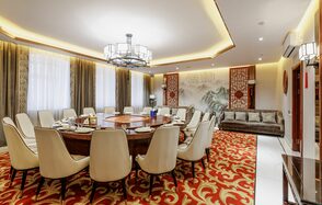 Ресторан J.Z.Peking Duck (J.Z. Утка по-пекински) . Просторная комната с круглым столом и белыми стульями, красный ковер с золотым узором, светлый потолок с подвесной люстрой, большие окна с занавесками, бежевый диван, китайская настенная роспись с текстом, декоративные панели по бокам.