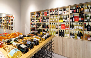 AVDRINKS (АВДРИНКС). Винный магазин с деревянными полками, полными бутылок вина, на двух уровнях. По центру — длинный стол с решетчатым основанием, на котором выложены бутылки. Освещение мягкое, с акцентом на стеллажи. Стены светлые, пол светлый.
