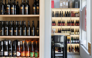 AVDRINKS (АВДРИНКС). Стеллажи с бутылками вина на деревянных полках, высокие стулья рядом с барной стойкой, современное освещение, светлые стены, витрина справа.