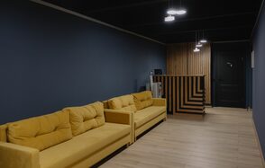 Танцевальный зал 65м2 студии RedHeartDance, Зал №1. Комната с двумя жёлтыми диванами вдоль тёмно-синей стены. Напротив них деревянная стойка с вертикальными полосами. На потолке висят подвесные светильники. Пол светло-бежевый, дверь тёмно-серая.