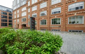Green Loft (Зеленый Лофт). Красное кирпичное здание с большими окнами. Перед ним тротуар из серых плит и зеленые кусты. У входа вертикальная черная вывеска с текстом. На первом этаже видна надпись «GREEN VKUS».