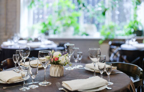 Green Loft (Зеленый Лофт). Круглый стол, сервированный белыми тарелками, стеклянными бокалами и салфетками. На столе цветочная композиция. Вокруг стулья темного цвета. На заднем плане большое окно с зелеными растениями. Естественное освещение.
