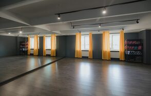 Танцевальный зал 50м2 студии RedHeartDance. Зал №3. Просторное помещение с зеркальными стенами, серыми стенами и полом из темного дерева. Большие окна с желтыми шторами. Черные трековые светильники на белом потолке. По бокам стеллажи с гимнастическими ковриками.