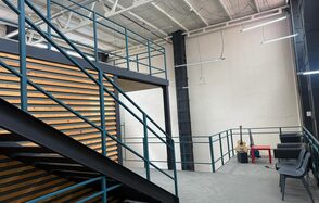 Просторный светлый лофт для мероприятий . Лофт-пространство с металлической лестницей и перилами, деревянная декоративная стенка, высокий потолок с подвесными светильниками. Серые диваны и стулья, красный стол. Светлые стены и бетонный пол.
